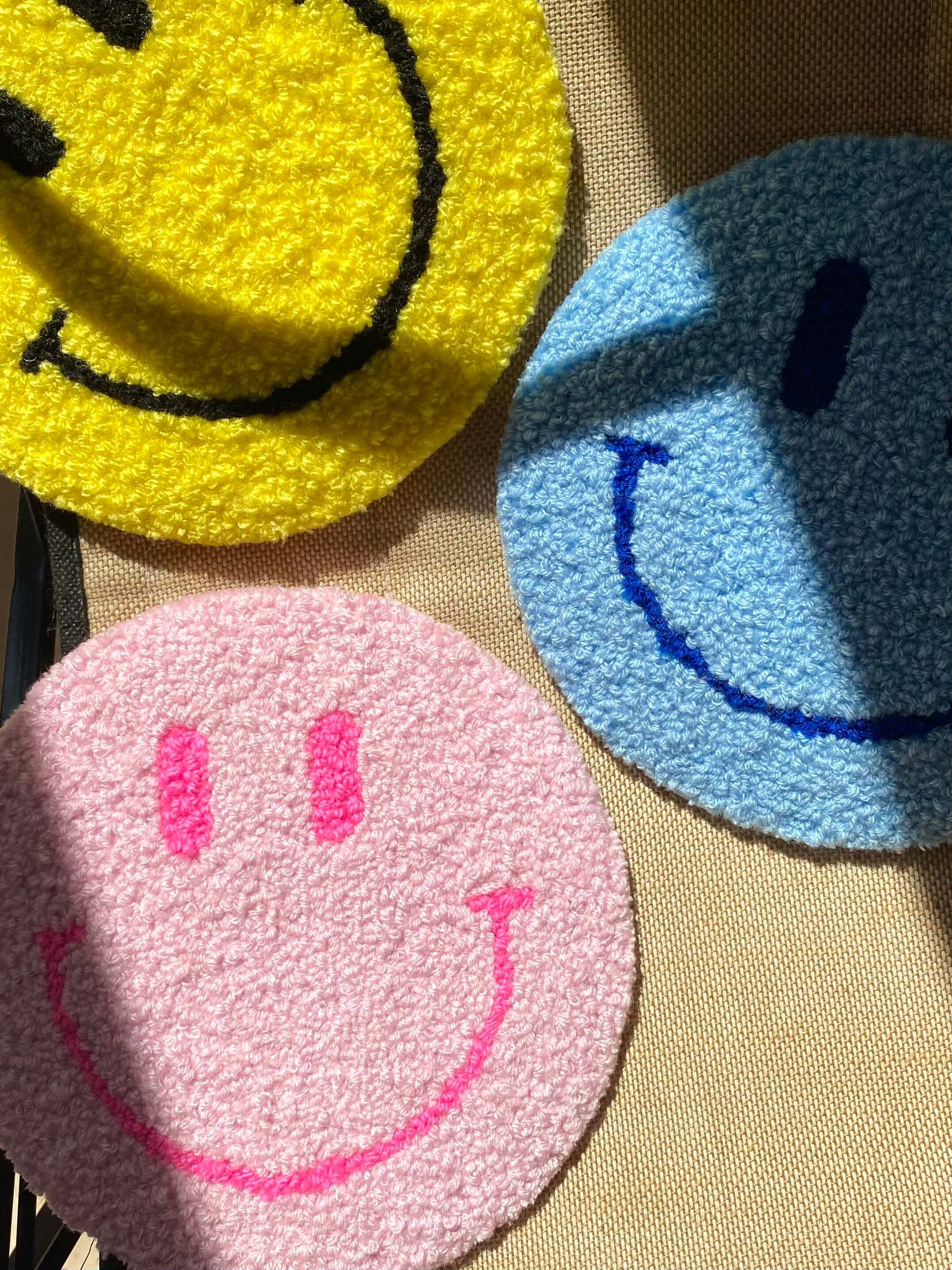 Tre Smiley vægtæpper i gul, blå og lyserød – lagt sammen på tekstiloverflade med sollys.