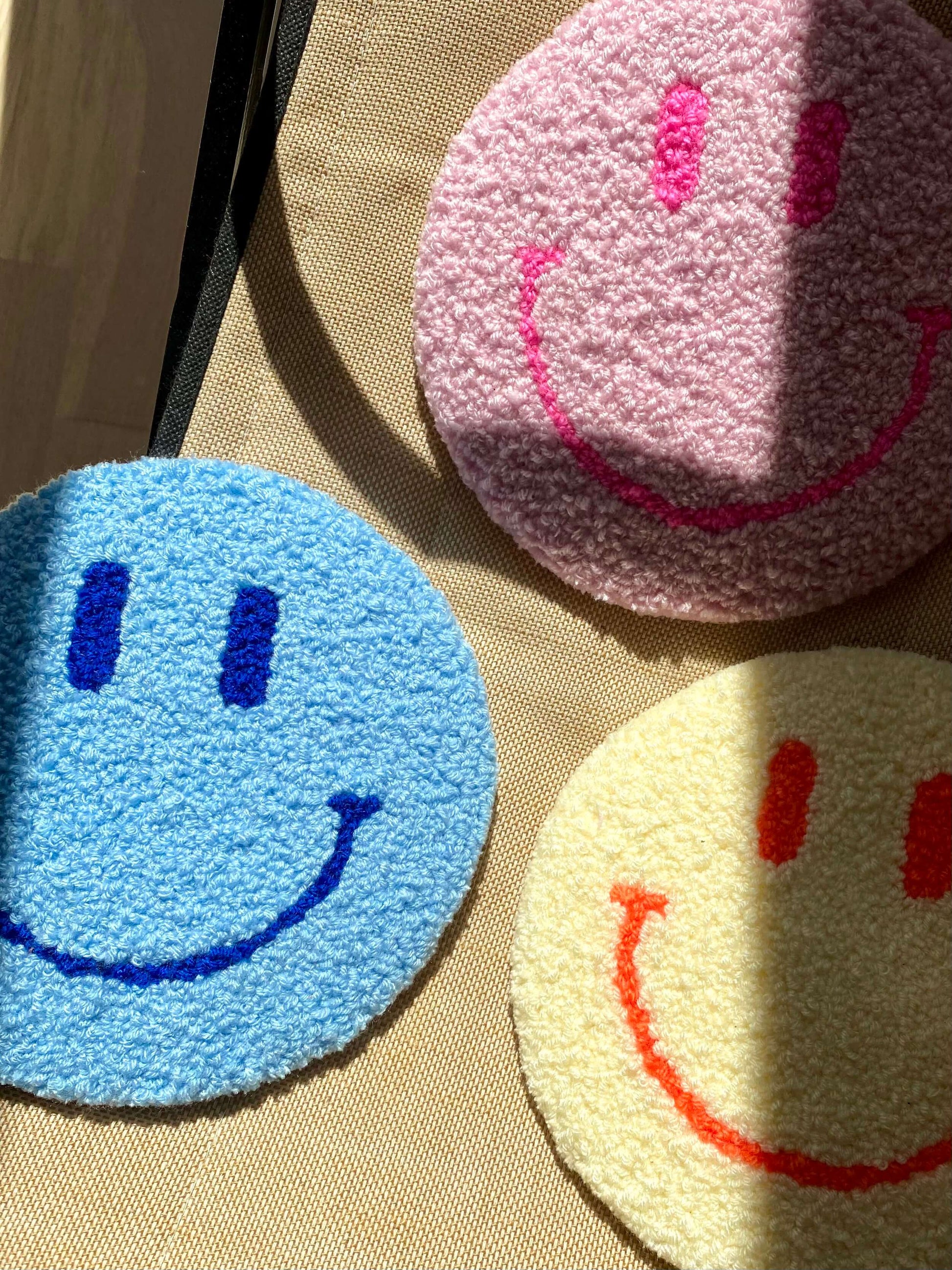 Tre Smiley vægtæpper i gul, blå og lyserød – lagt sammen på tekstiloverflade med sollys.