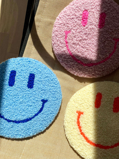Tre Smiley vægtæpper i gul, blå og lyserød – lagt sammen på tekstiloverflade med sollys.