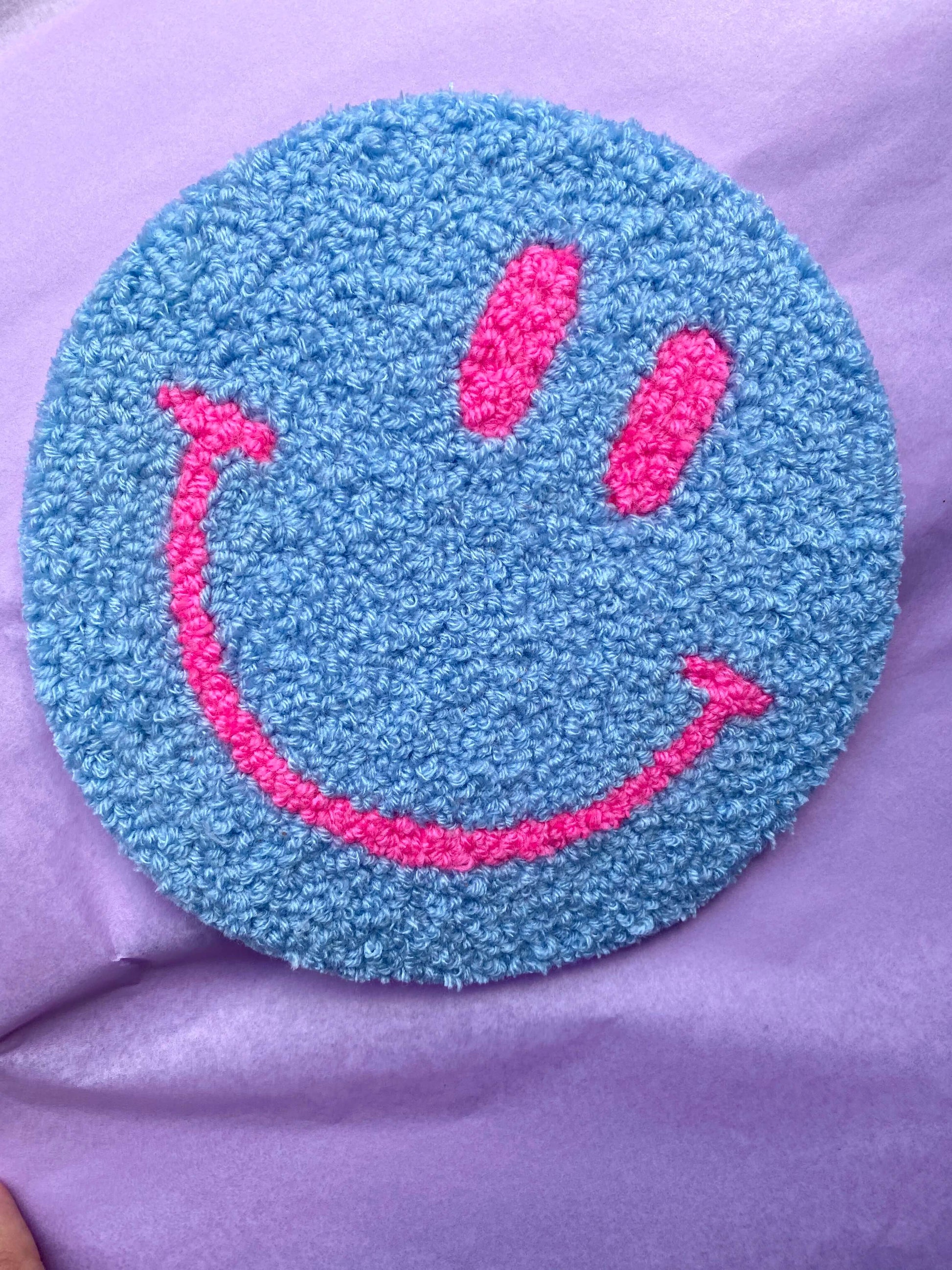 Blå Smiley vægtæppe med pink ansigtstræk – håndlavet vægtæppe på lilla baggrund.